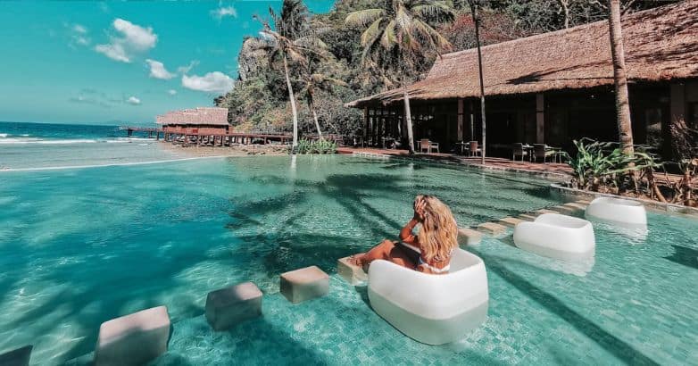 Woman on a floating seat in a tropical pool, surrounded by palm trees, villas, and a stunning ocean view."