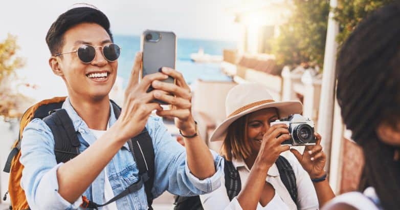 Happy travelers capturing moments with a smartphone and camera, illustrating how travel content marketing can leverage social media to engage audiences and showcase authentic experiences.